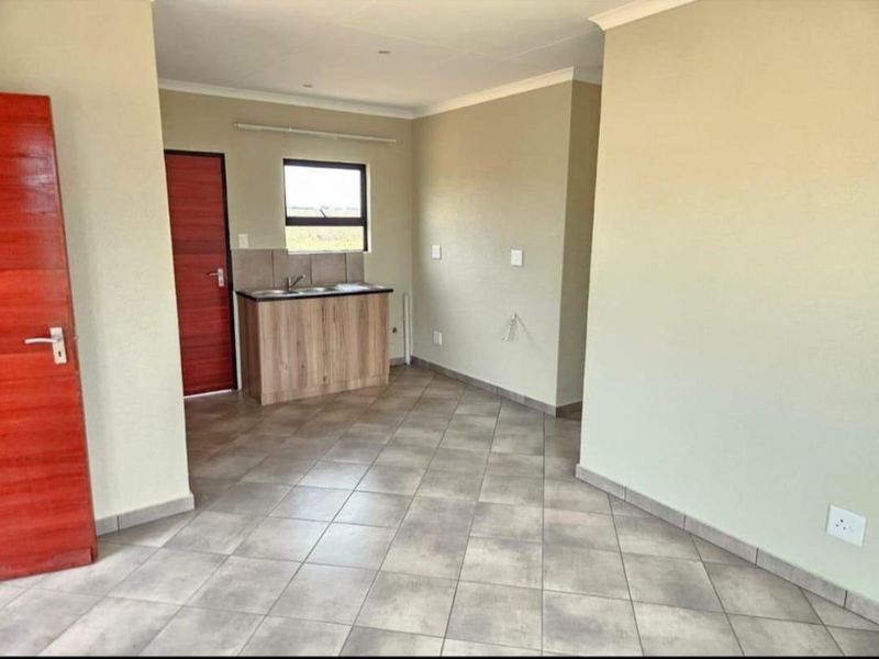 An open-plan living area featuring tiled flooring, neutral walls, and a compact kitchenette with built-in cabinetry and a sink.
