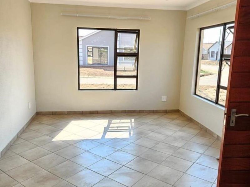 A bright, empty living area featuring neutral walls, tiled flooring, and large aluminium-framed windows providing natural light.