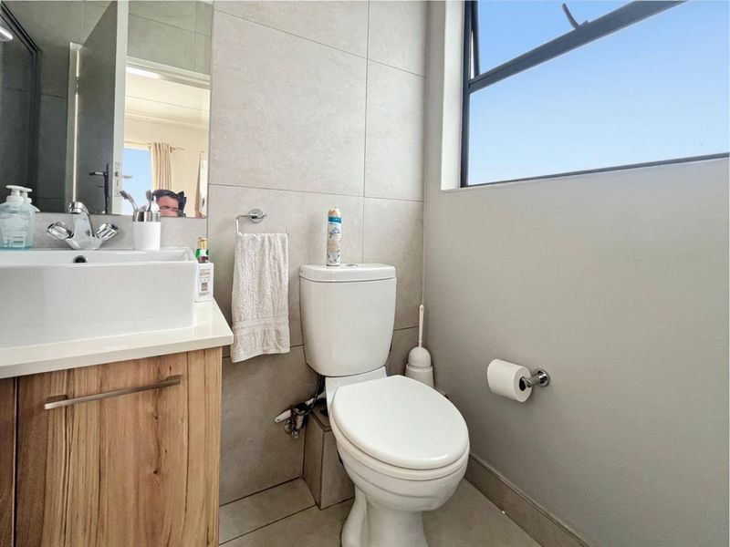 A modern bathroom featuring a wood-finish vanity cabinet, ceramic basin, wall-mounted mirror, and tiled finishes with natural light from a top-hung window.
