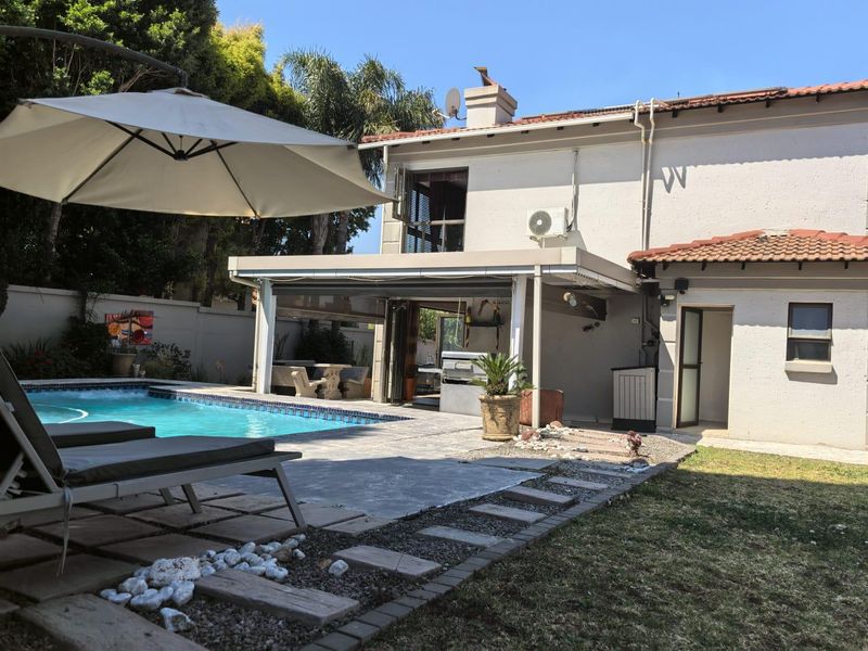 A well-maintained outdoor entertainment area featuring a swimming pool, covered patio, and established garden surroundings.