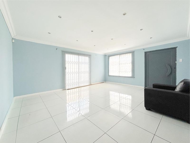 A bright and spacious living area featuring white tiled flooring, recessed downlights, and a sliding glass door with security gate access.