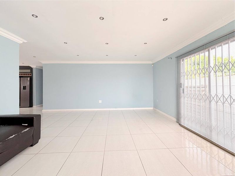 A spacious, light-filled living area featuring neutral tiled flooring, recessed downlighting, and a sliding glass door with integrated security gate.