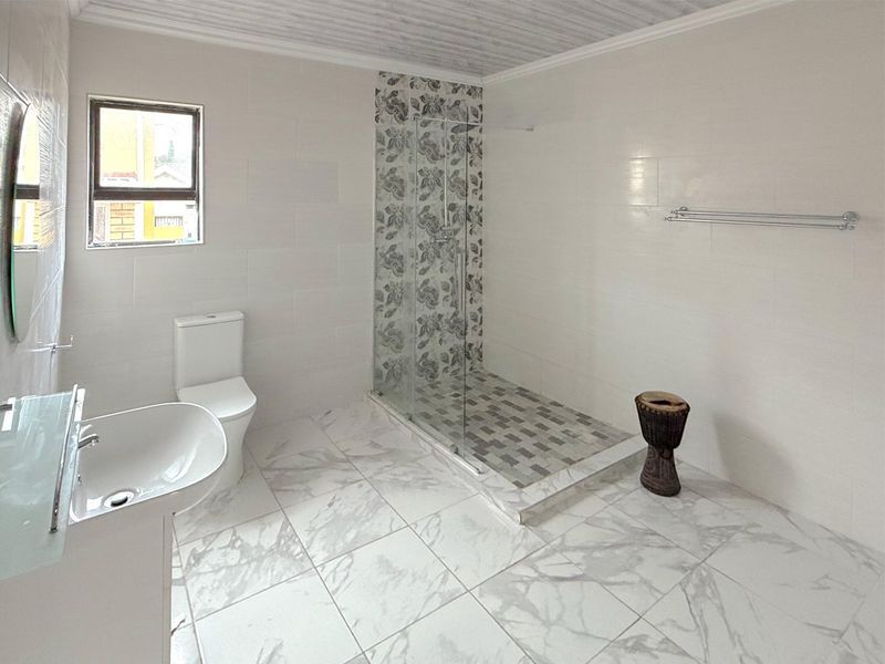 A modern, bright bathroom featuring a walk-in shower with decorative tiling, a pedestal basin, and neutral floor-to-ceiling wall tiles.
