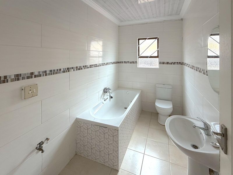 A clean, tiled bathroom featuring a bathtub, pedestal sink, and toilet, with neutral finishes and a window for natural light.