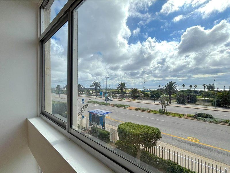 A clear view from an apartment window overlooking a coastal road, palm trees, and the ocean horizon.