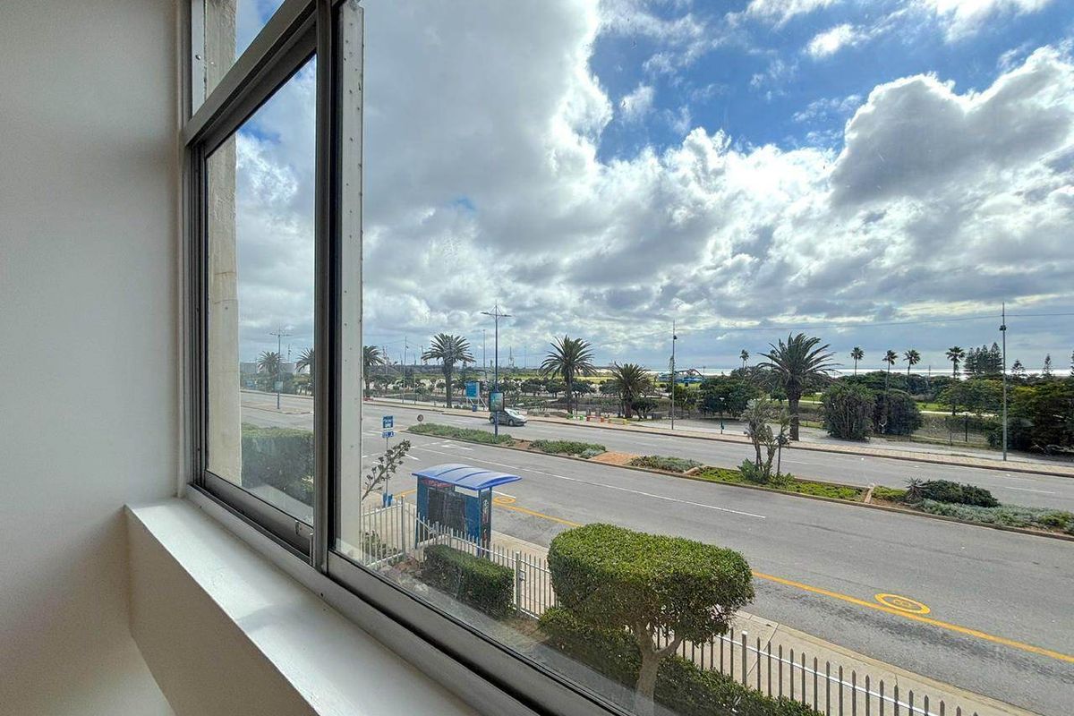 A clear view from an apartment window overlooking a coastal road, palm trees, and the ocean horizon.