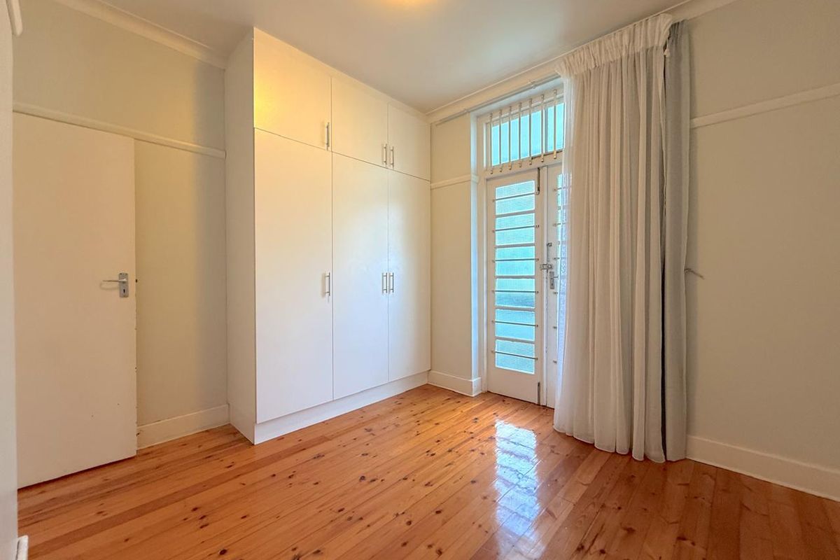 A bright, empty bedroom featuring polished timber flooring, ample built-in cupboard storage, and a glass-panelled door providing natural light.