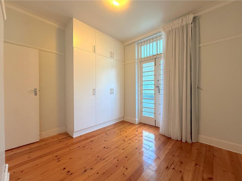 A bright, empty bedroom featuring polished timber flooring, ample built-in cupboard storage, and a glass-panelled door providing natural light.