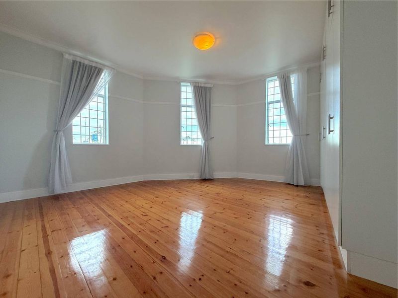 A bright, empty bedroom featuring polished timber flooring, built-in cupboards, and three large multi-pane windows allowing for ample natural light.