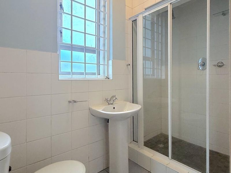 A functional bathroom featuring a pedestal basin, a glass-enclosed shower, and neutral wall and floor tiling.