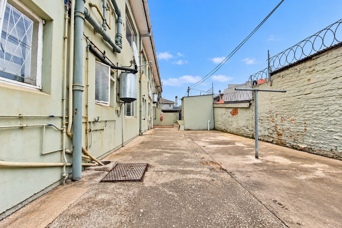 Rear exterior service area featuring concrete paving, boundary wall with razor wire, and exposed external plumbing infrastructure.