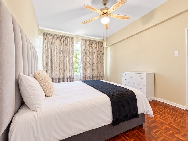 A well-maintained bedroom featuring classic parquet flooring, a central ceiling fan, and large windows dressed with curtains.