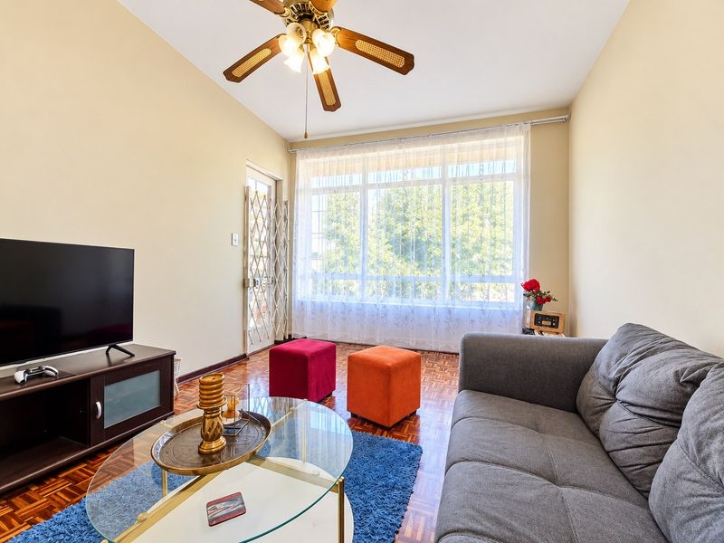 A bright living room featuring classic parquet flooring, a ceiling fan, and a large window with a security gate leading to the exterior.