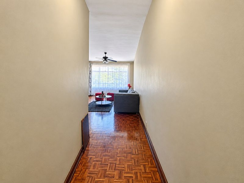 A view through a hallway with parquet flooring leading into a living area with a ceiling fan and large window.