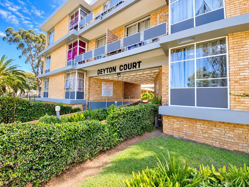 Exterior view of Deyton Court apartment building featuring a brick facade, private balconies with metal railings, and a landscaped front garden area.