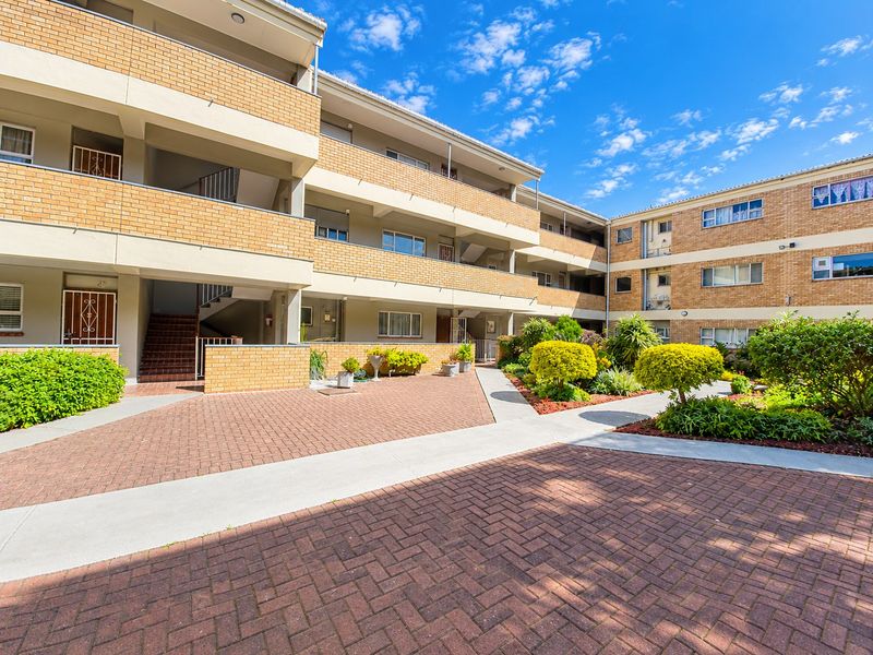 A well-maintained apartment complex exterior featuring a brick facade, paved courtyard, and landscaped garden areas with accessible staircases and balconies.