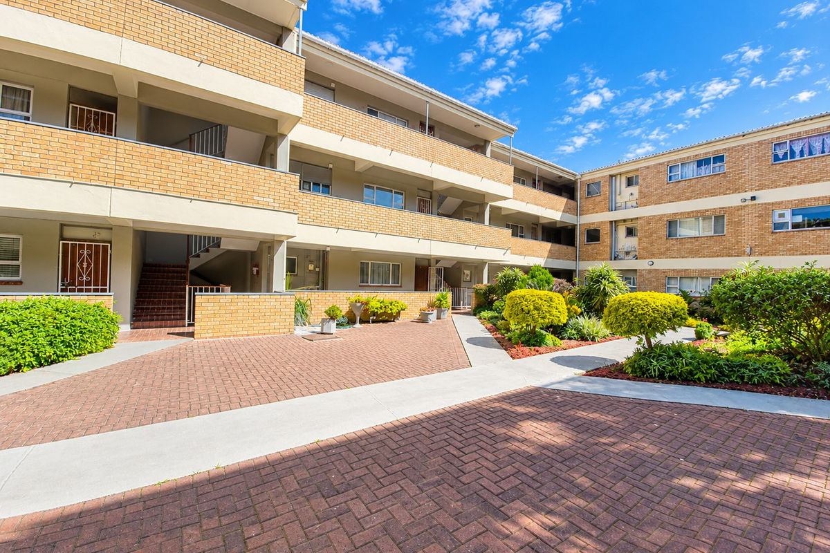A well-maintained apartment complex exterior featuring a brick facade, paved courtyard, and landscaped garden areas with accessible staircases and balconies.