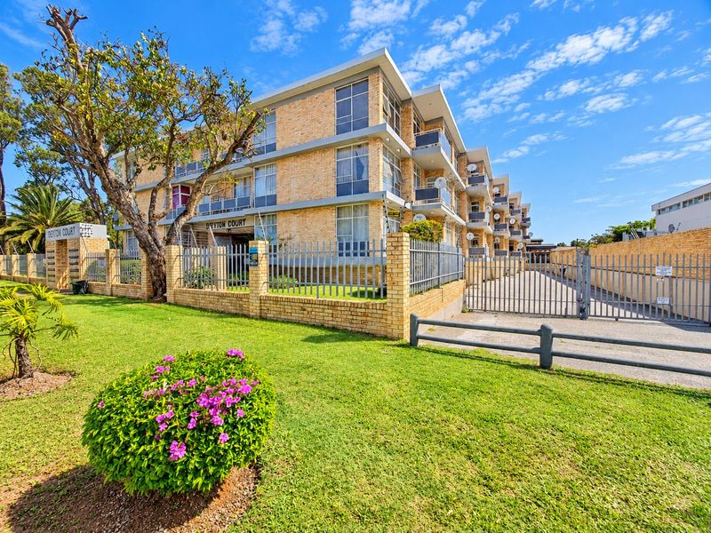 Exterior view of Deyton Court apartment complex featuring a brick facade, private balconies, and secure gated driveway access.