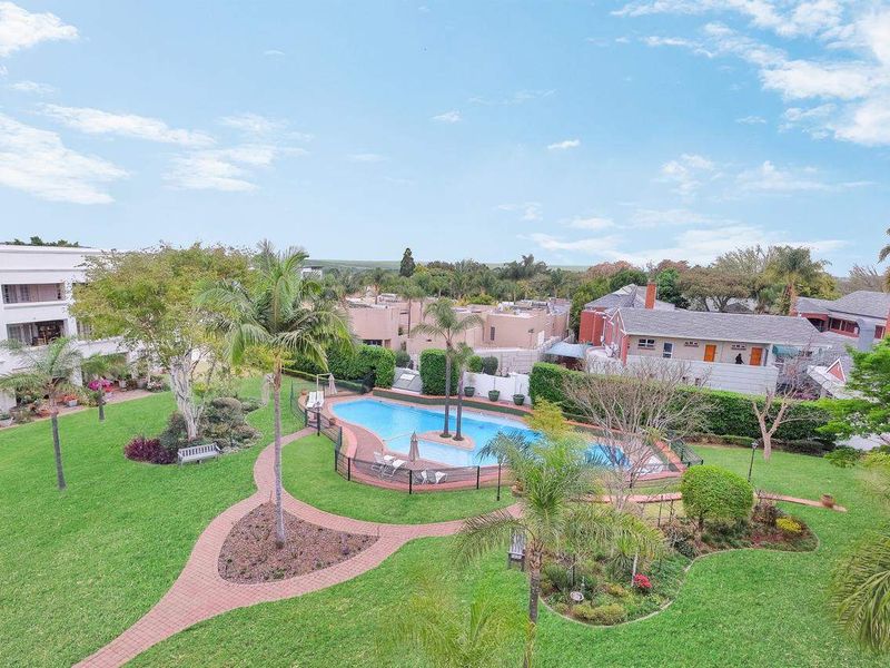An elevated aerial view of the apartment complex grounds, featuring a well-maintained swimming pool, lush landscaped gardens, and paved pathways.