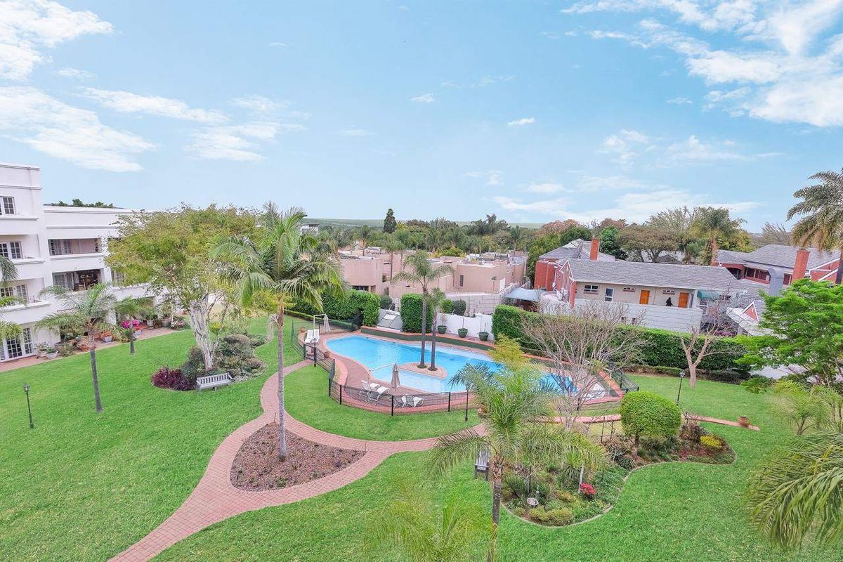 An elevated aerial view of the apartment complex grounds, featuring a well-maintained swimming pool, lush landscaped gardens, and paved pathways.