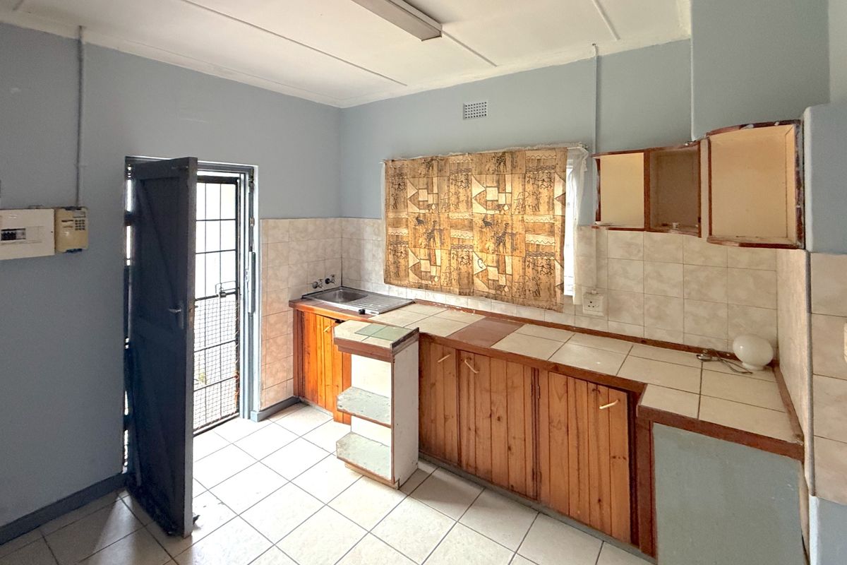 A dated kitchen featuring wooden cabinetry, tiled countertops, and a stainless steel sink, with direct access to an exterior security door.