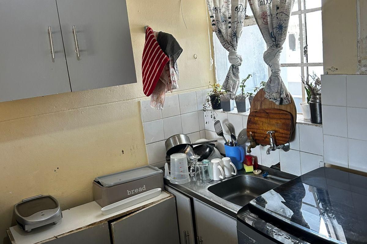 A compact kitchen area featuring grey cabinetry, a stainless steel sink, and tiled splashbacks, offering a functional layout in need of cosmetic updates.