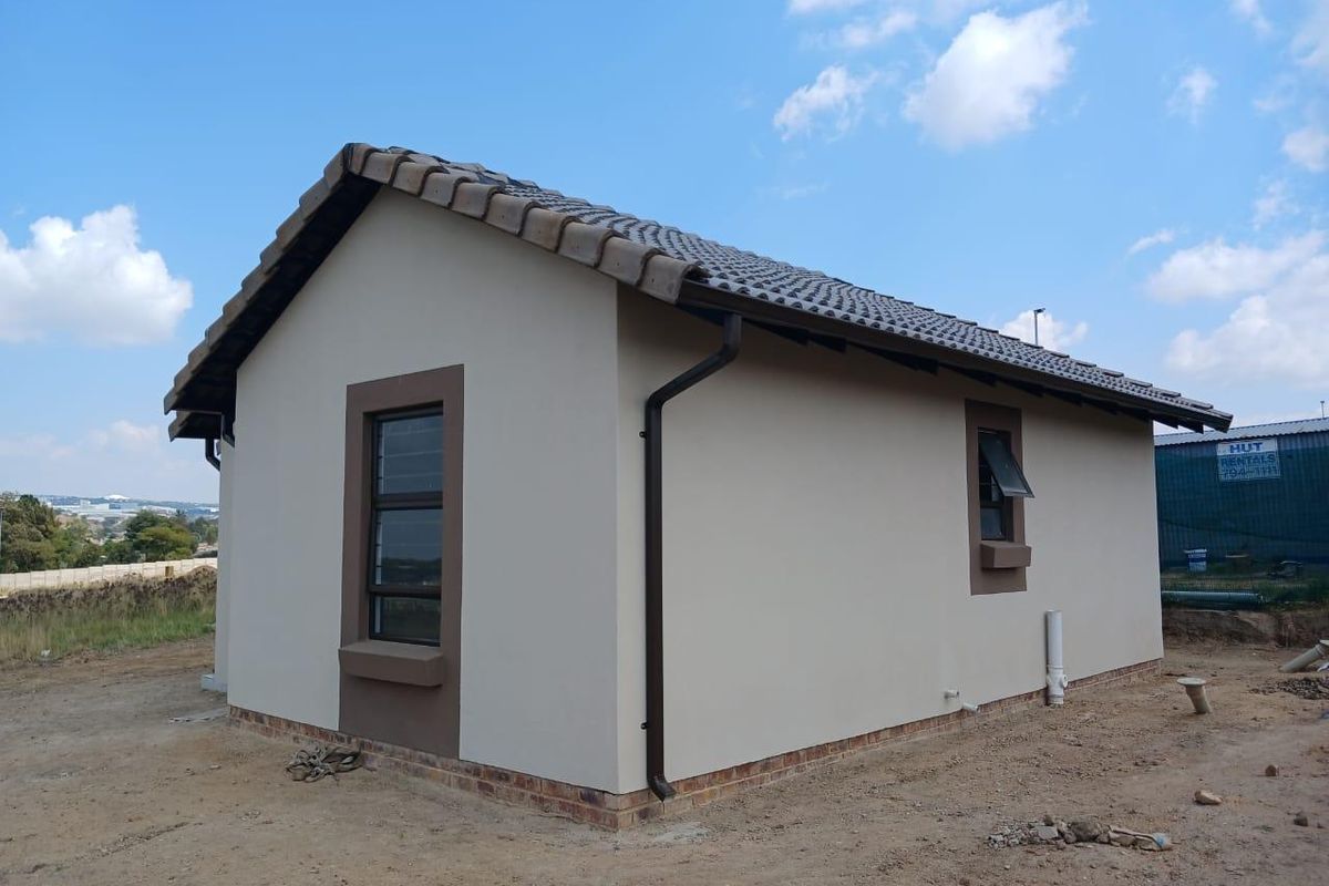 Exterior view of a modern single-story house featuring a tiled roof, painted plaster walls, and aluminum window frames, set on an undeveloped plot.