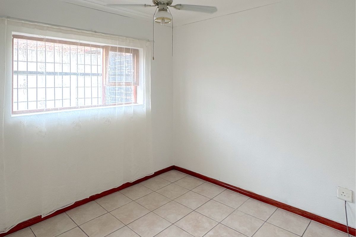 A bright, empty bedroom featuring neutral tiled flooring, a ceiling fan, and a window fitted with security bars.