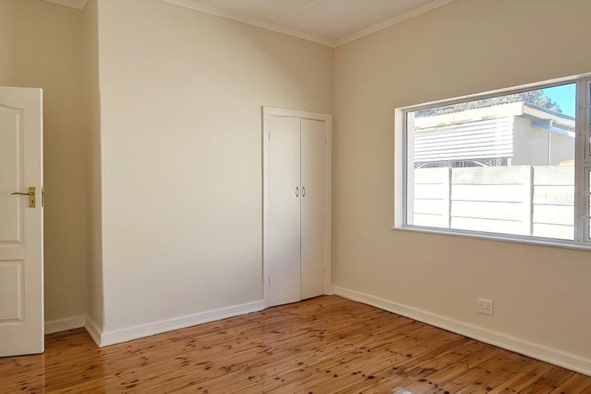 A bright, empty bedroom featuring polished wooden flooring, built-in cupboards, and a large window with security bars.