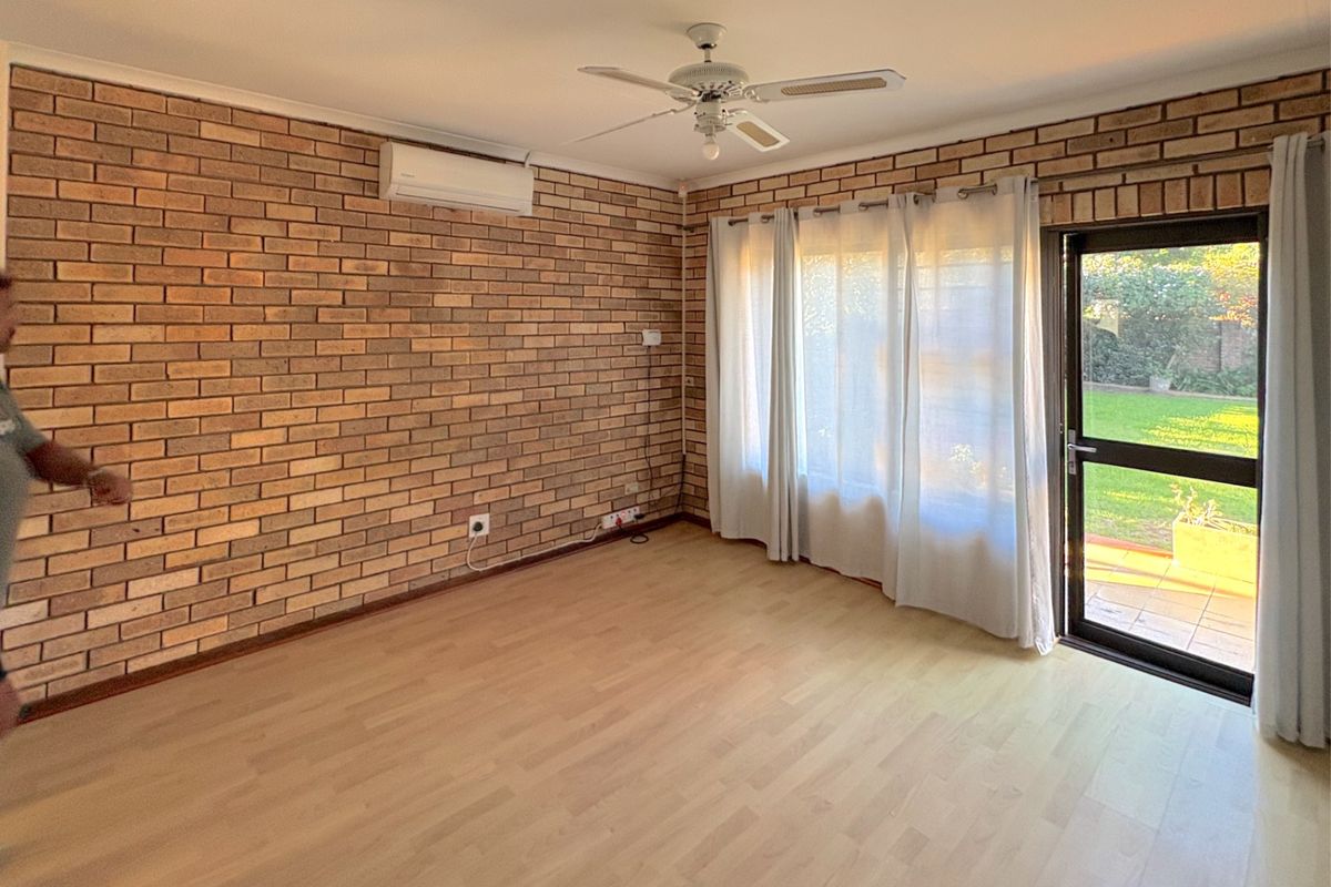A living room featuring a prominent face-brick feature wall, light wood-look flooring, a ceiling fan, and a split-system air conditioning unit with garden access.