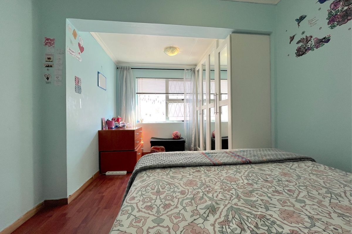 A bedroom featuring built-in mirrored wardrobes, laminate flooring, and a window with blinds, providing a functional and well-lit sleeping space.
