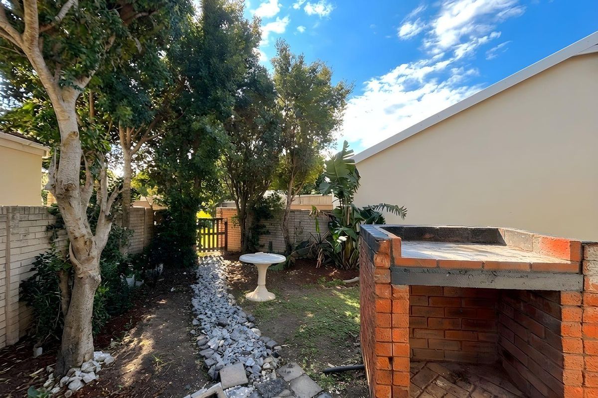 A private rear garden featuring a built-in brick braai, mature trees, and a stone-lined pathway leading to a wooden gate.
