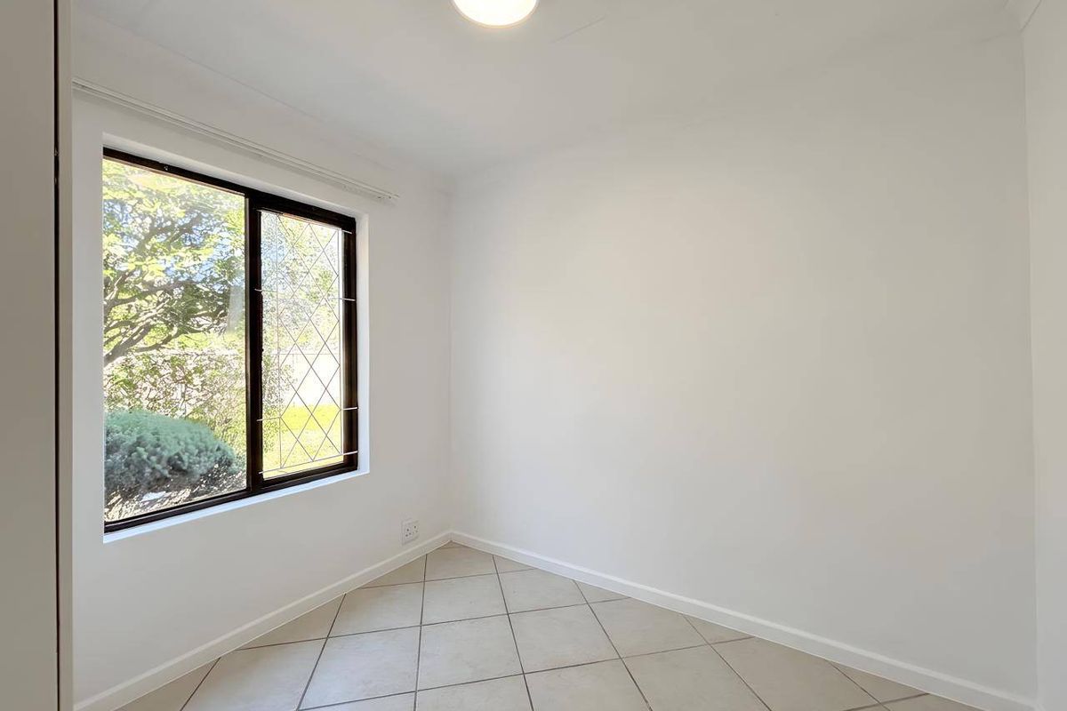 A bright, empty bedroom featuring neutral tiled flooring, a large window with security bars, and a simple ceiling light fixture.