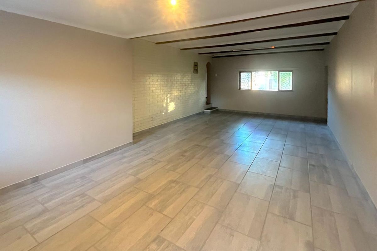 A spacious, empty living area featuring tiled flooring, exposed ceiling beams, and a painted brick feature wall with natural light from a window.