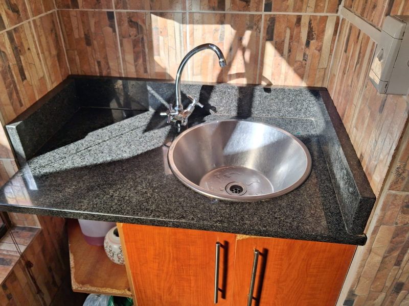 A compact utility area featuring a granite countertop, stainless steel basin, and wooden cabinetry with tiled wall finishes.