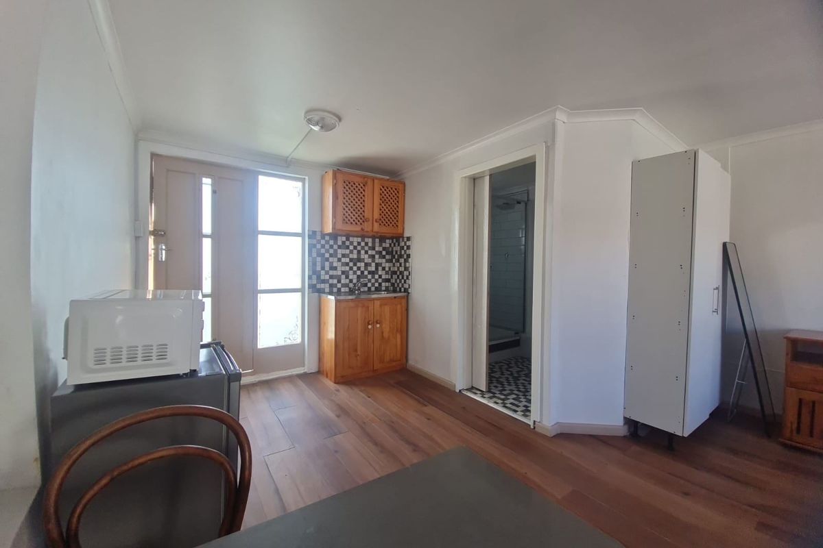 A compact kitchenette area featuring wooden cabinetry, a tiled splashback, and direct access to an adjoining bathroom and exterior door.