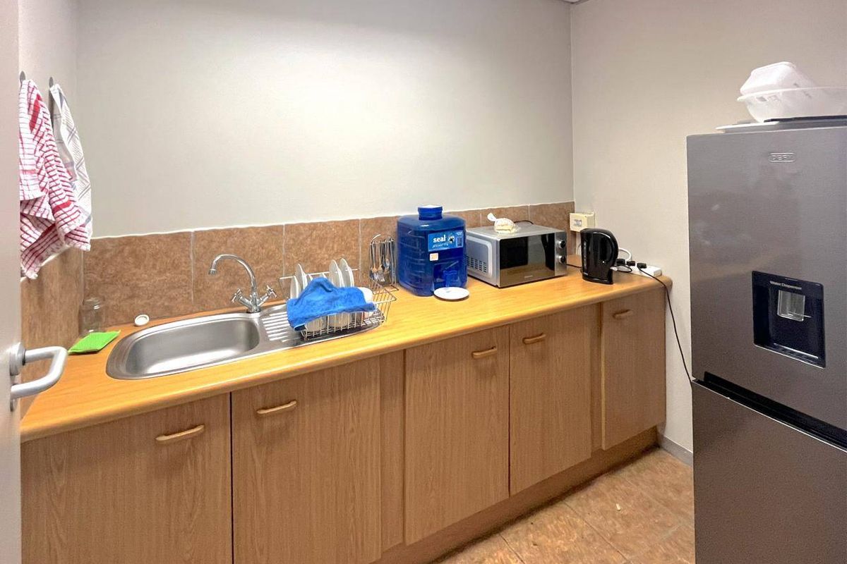 A functional kitchenette area featuring wood-look cabinetry, a stainless steel sink, tiled splashback, and space for a refrigerator.