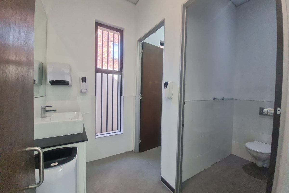 A clean commercial restroom featuring a modern basin, wall-mounted soap dispenser, and a private toilet cubicle with tiled walls and secure window bars.
