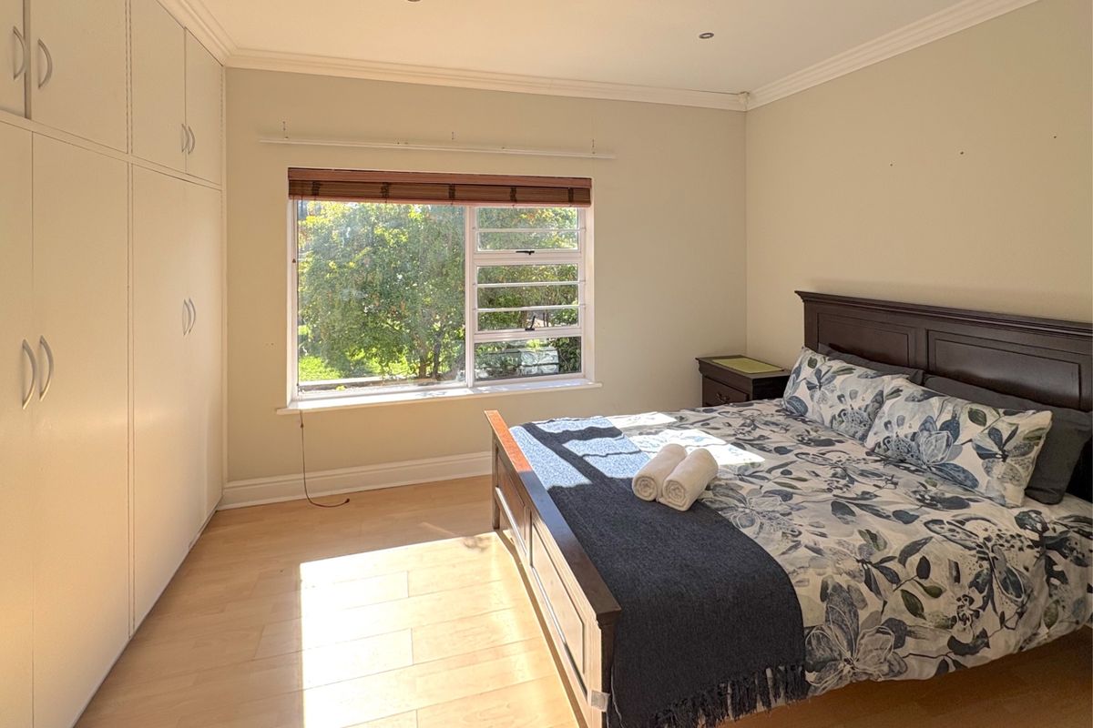 A bright bedroom featuring light wood-look flooring, built-in floor-to-ceiling wardrobes, and a large window with garden views.
