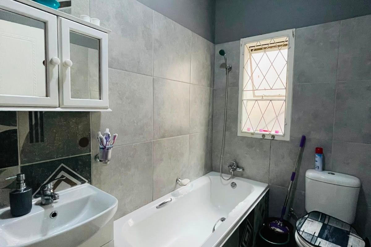 A functional bathroom featuring a built-in bathtub, wall-mounted vanity with basin, and a toilet, finished with grey wall tiles and a high ceiling.