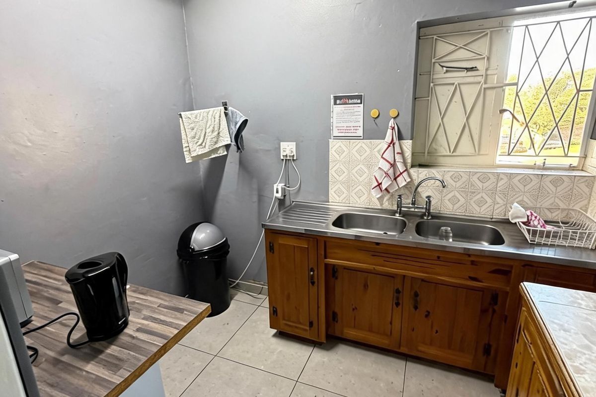 A functional kitchen area featuring wooden cabinetry, a double stainless steel sink, tiled flooring, and a window with security bars.