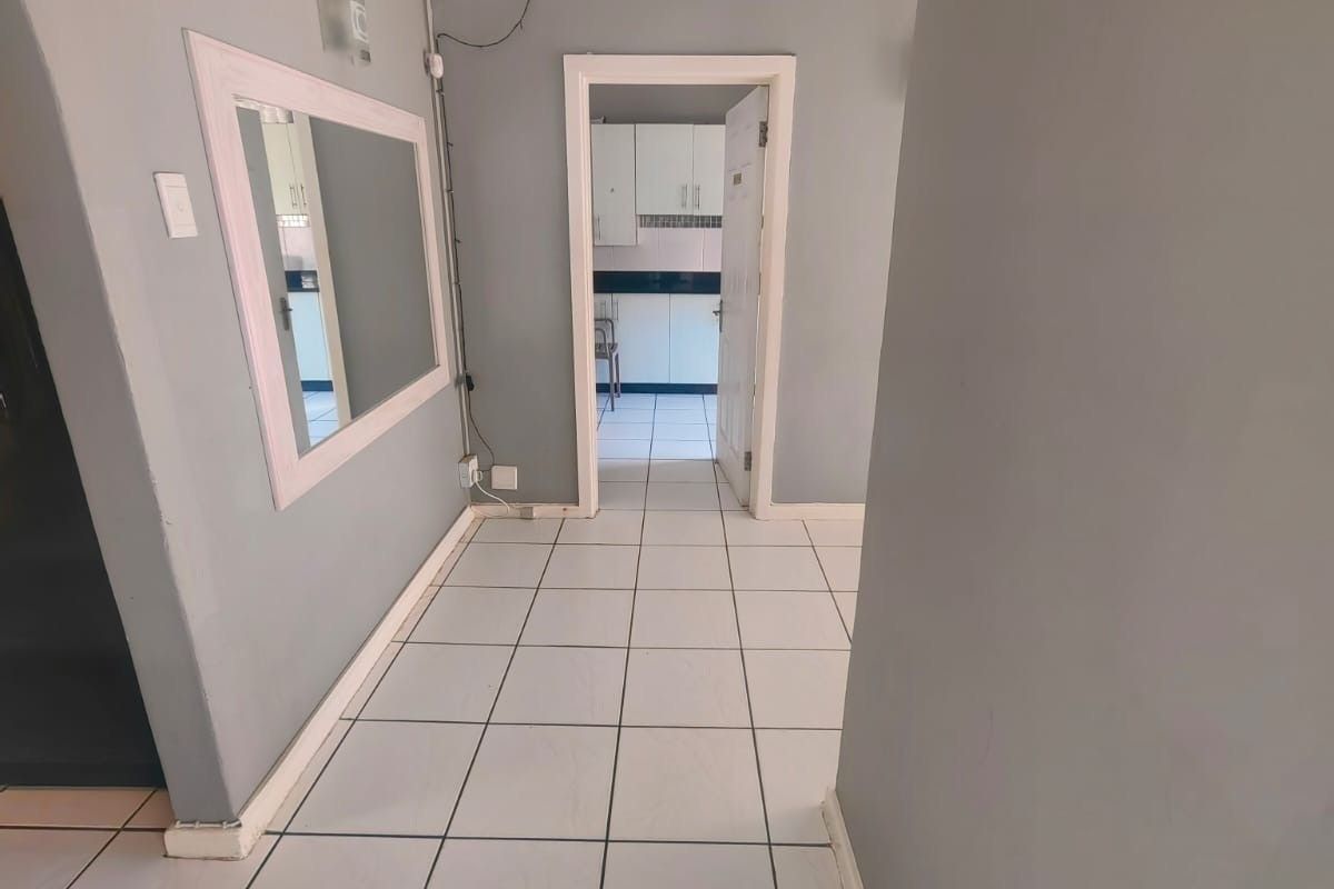 A neutral-toned hallway featuring ceramic floor tiles, coved ceiling detailing, and a wall-mounted mirror, providing access to an adjacent kitchen area.