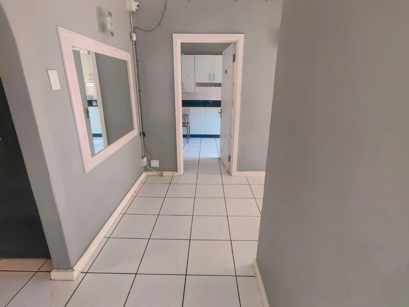 A neutral-toned hallway featuring ceramic floor tiles, coved ceiling detailing, and a wall-mounted mirror, providing access to an adjacent kitchen area.