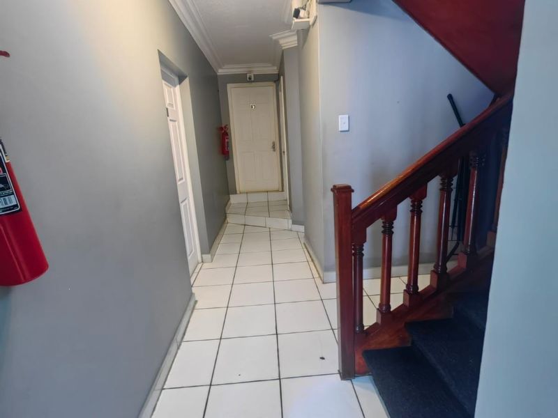 A commercial hallway featuring white tiled flooring, a wooden staircase with a balustrade, and wall-mounted fire safety equipment.