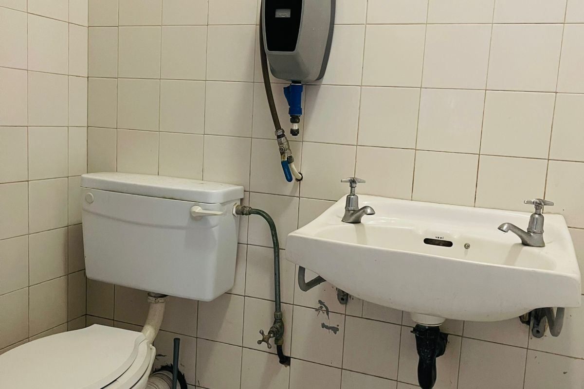 A dated bathroom featuring a wall-mounted basin, toilet, and electric water heater with visible plumbing and worn wall and floor tiling.