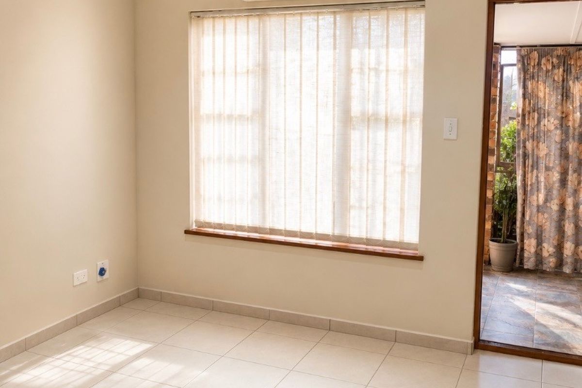 A bright, empty bedroom featuring neutral tiled flooring, recessed ceiling lighting, a wall-mounted air conditioning unit, and a window with vertical blinds.