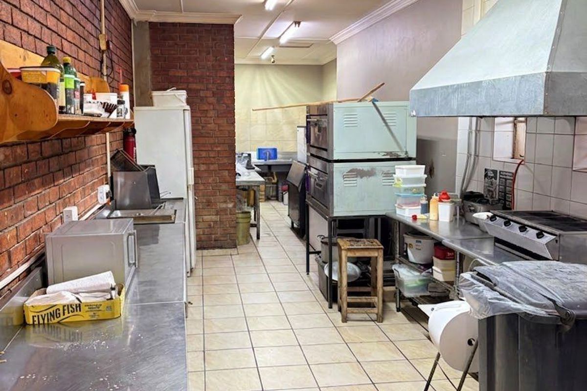 Commercial kitchen featuring stainless steel prep counters, industrial extractor hood, tiled flooring, and exposed brick wall detailing.