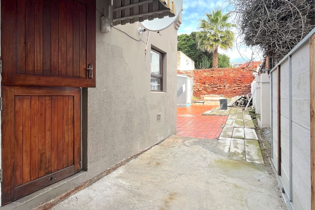 A side exterior view of the property featuring a stable-style wooden door, concrete pathway, and a rear paved area with a brick boundary wall.