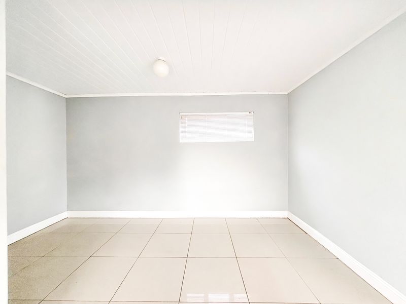 A bright, empty bedroom featuring neutral tiled flooring, light grey walls, and a tongue and groove ceiling with a central light fixture.
