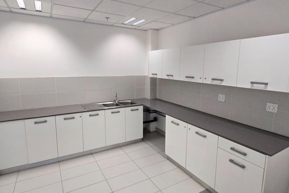 A clean, functional commercial kitchenette featuring white cabinetry, grey countertops, tiled splashbacks, and a stainless steel sink.